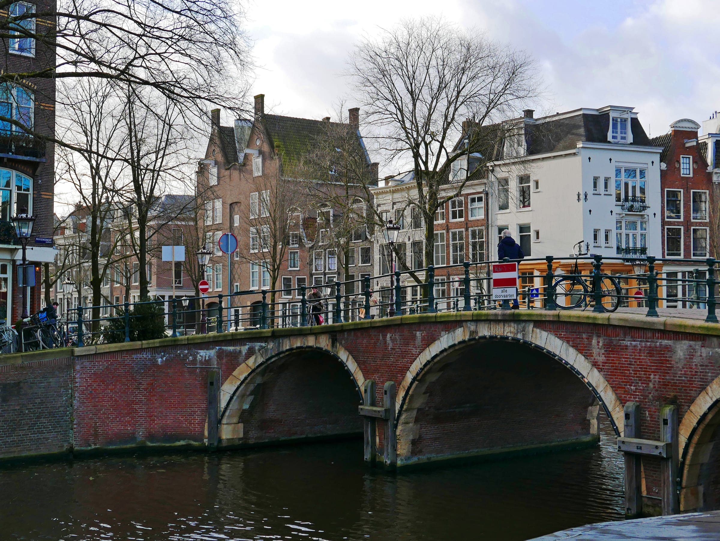 A red-brick arched bridge over an Amsterdam canal, with bicycles and a person leaning on its iron railing, and rows of gabled canal houses and bare trees behind it.