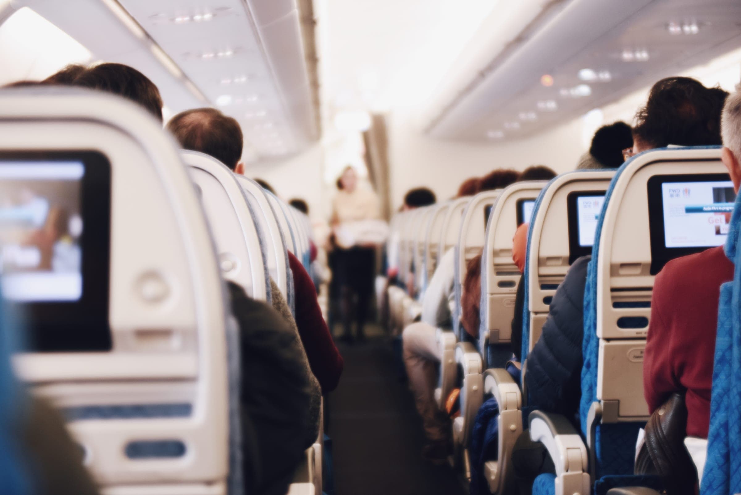 A cabin crew member in a pale uniform stands at the far end of an airliner's aisle during a flight, seen past rows of seated passengers from behind and lit seatback screens on either side.