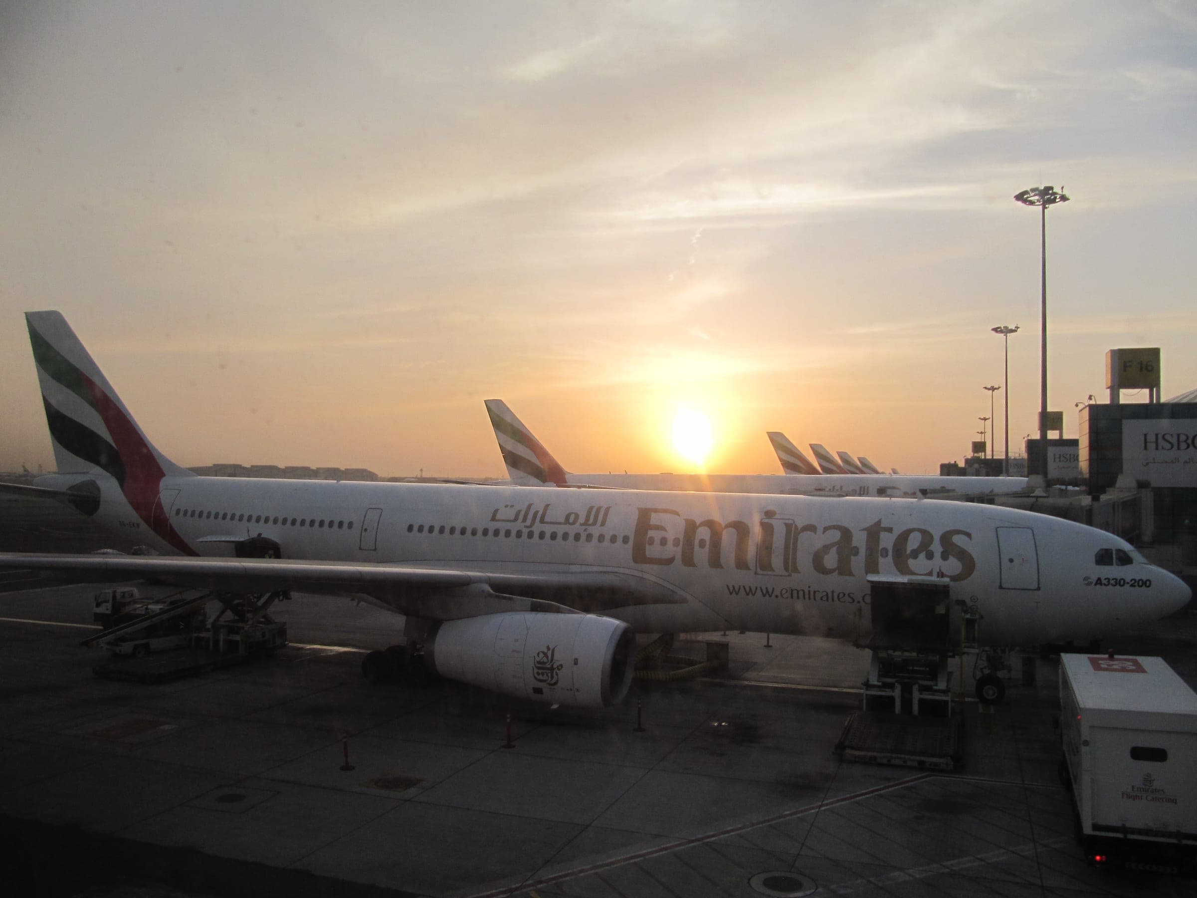 A line of Emirates Airbus A330s parked at their gates at Dubai International as the sun sets behind the terminal.