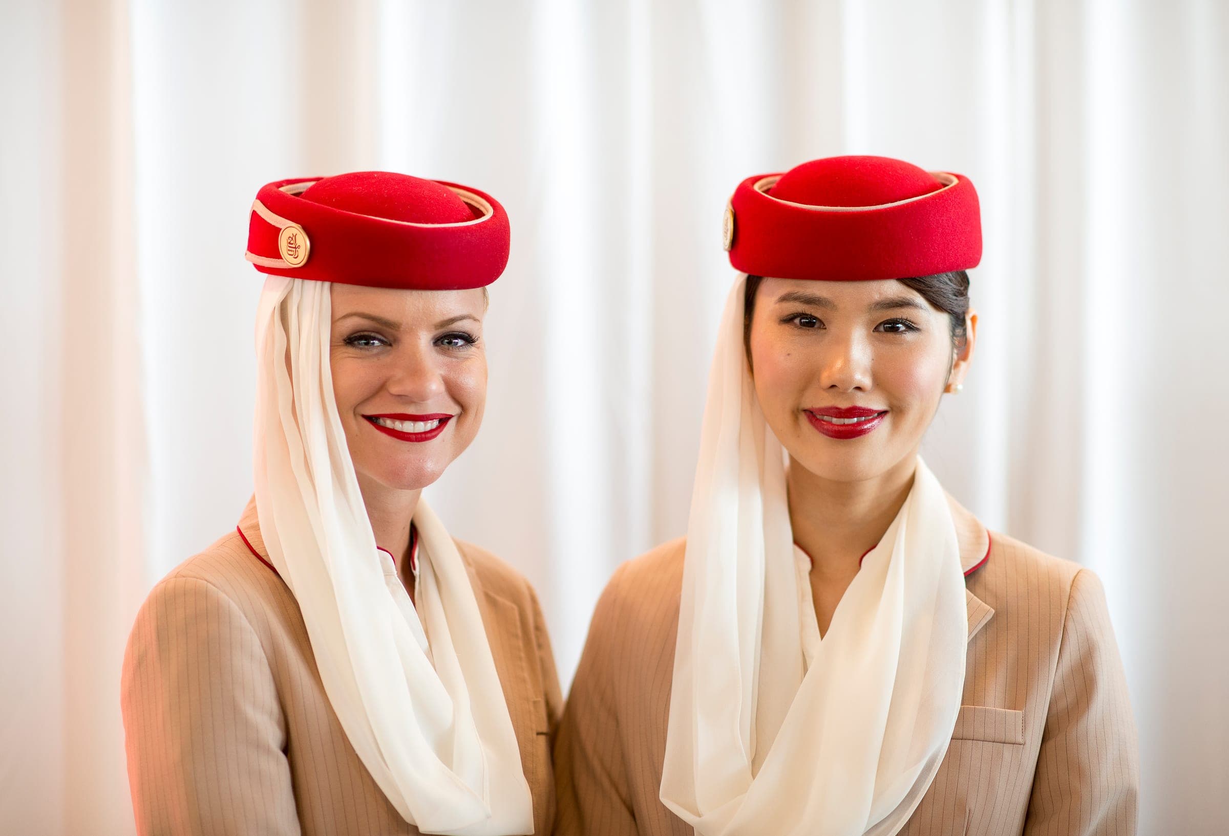 Two Emirates cabin crew members in the airline's uniform, a beige jacket with a red hat and white veil, smiling at the camera at Brussels Airport.