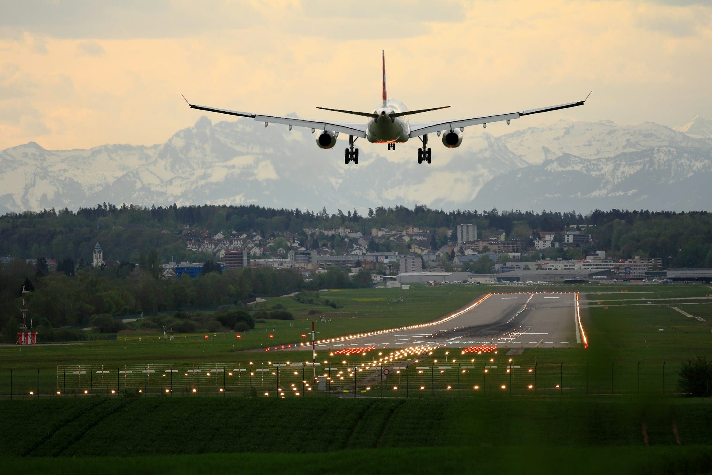 A twin-aisle airliner on short final over a lit approach path, snow-covered alpine peaks rising behind the airport.
