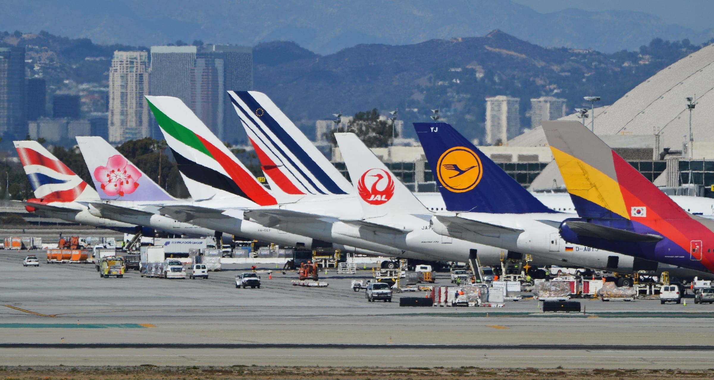 The tails of seven long-haul airliners parked side by side at Los Angeles International Airport, from left British Airways, China Airlines, Emirates, Air France, Japan Airlines, Lufthansa and Asiana Airlines, with ground vehicles on the apron below.