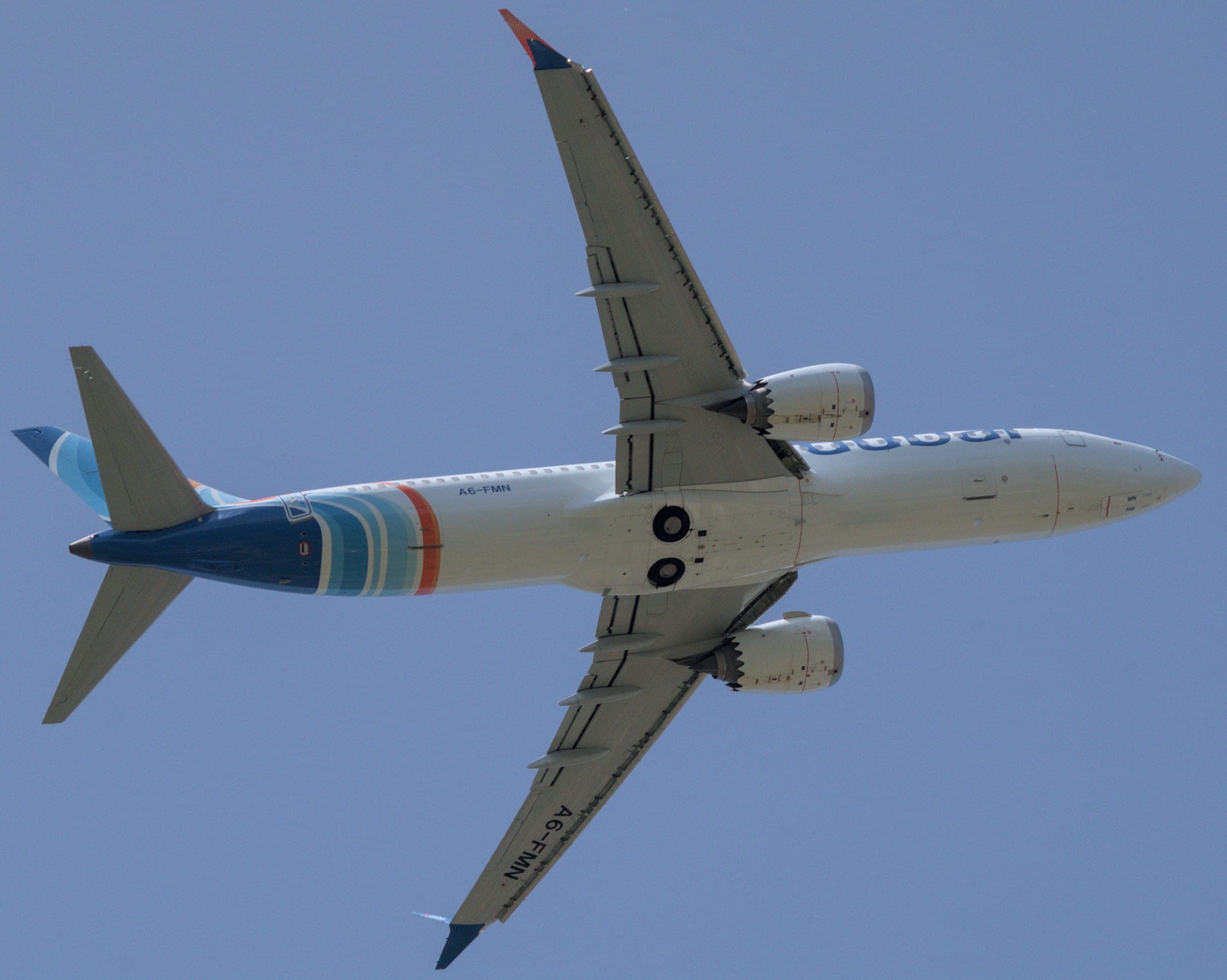 A flydubai Boeing 737 MAX 8, registration A6-FMN, seen from directly below against a clear blue sky as it takes off from Pisa, in white with blue, turquoise and orange bands towards the tail.