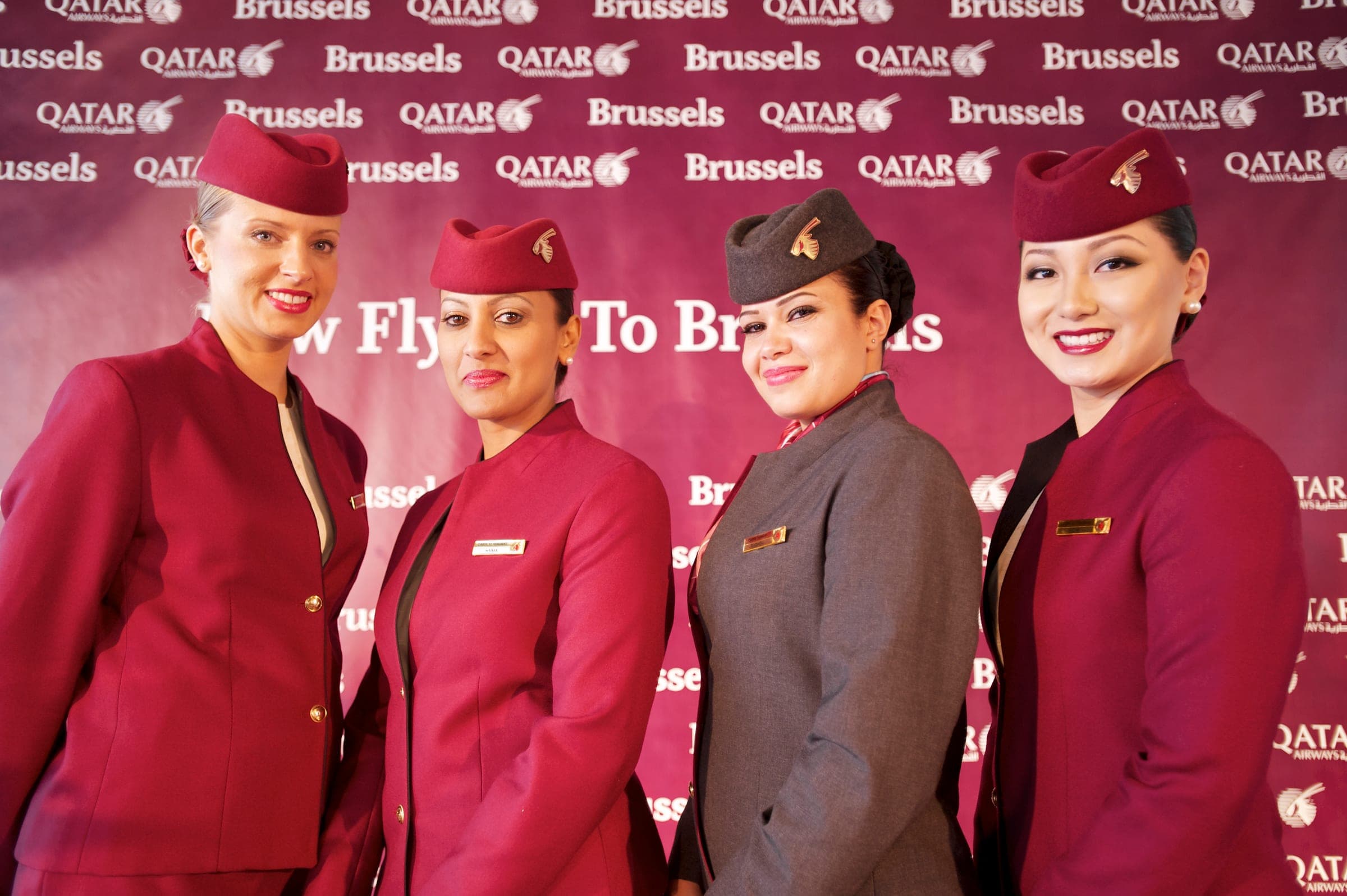 Four Qatar Airways cabin crew members, three in the burgundy uniform and hat and one in grey, smiling in front of a Qatar Airways and Brussels backdrop at Brussels Airport in 2011.