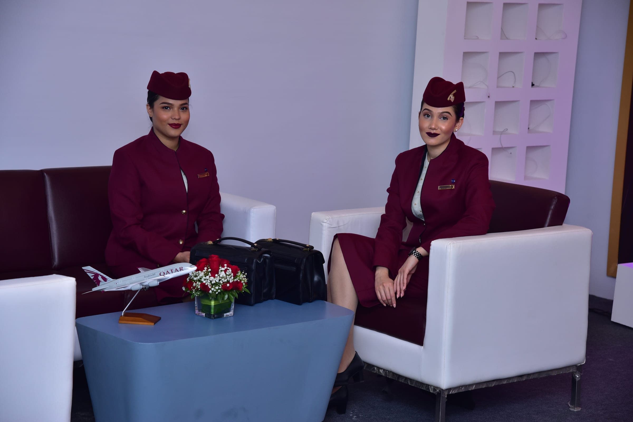 Two Qatar Airways cabin crew in the airline's burgundy uniform and hats sit on a white sofa beside a table holding a model Qatar Airways A380 and a vase of red roses.