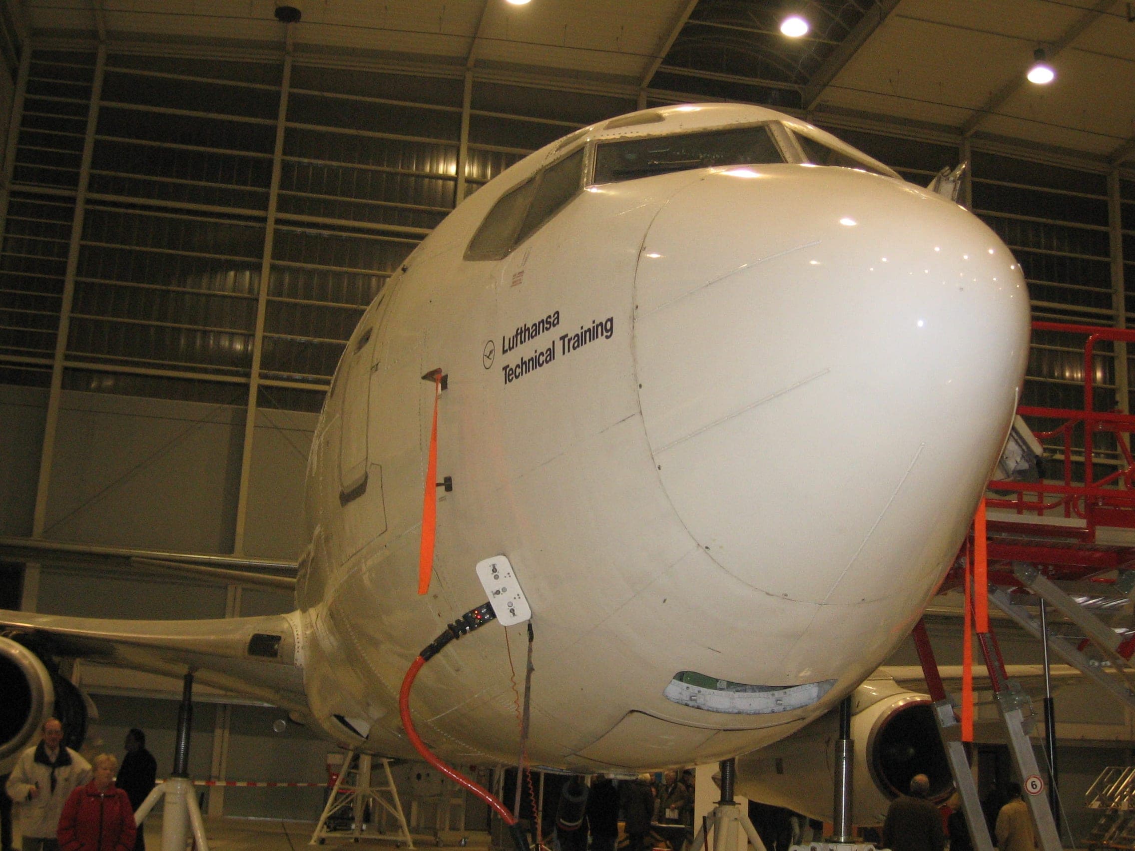 The nose of a Boeing 737 inside a maintenance hangar, lettered Lufthansa Technical Training, with red access platforms drawn up alongside.